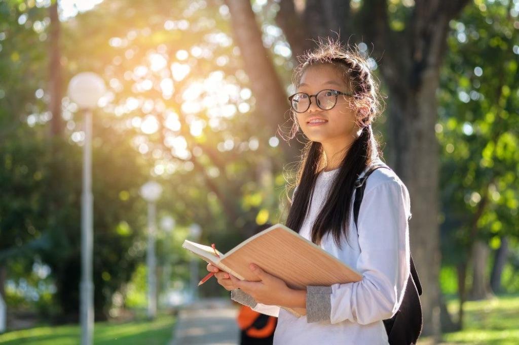 junior asian girls student with glasses are standing reading a book in garden t20 0AY76k