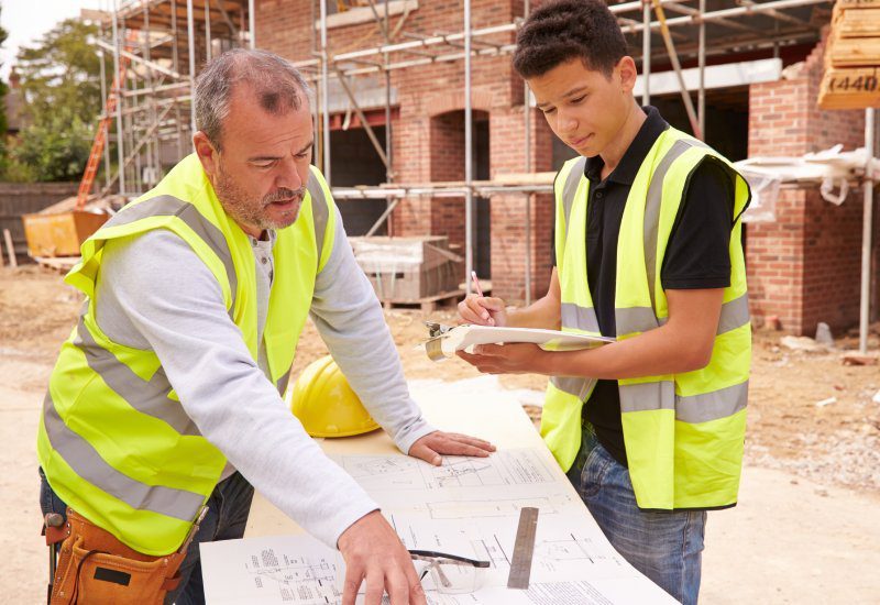 older construction worker explaining building plans to a teenage apprentice on a construction site