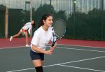 Two teen girls playing tennis. The girl in front is holding up her racquet. The girl in the back is returning the ball.