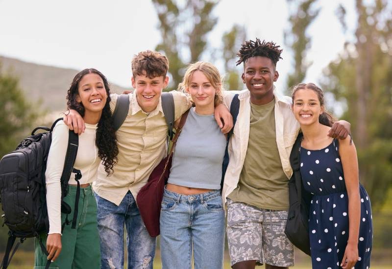 Three young women and two young men stand with their arms around each other, all wearing backpacks and smiling
