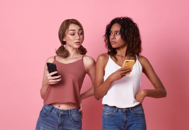 Two young women stand next to each other holding smartphones. One of them peeks over the other's shoulder to read her screen.