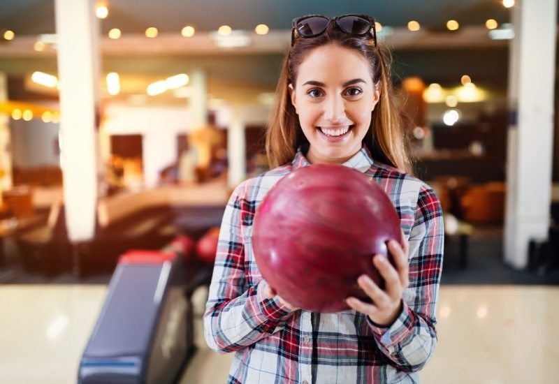 A teenage girl wearing flannel in a bowling alley, smiling and holding a red bowling ball at chest level.