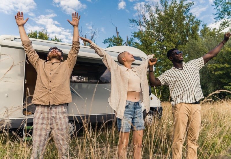 A trio of friends standing outside of their white van, holding their arms out to the air as they stretch and smile.