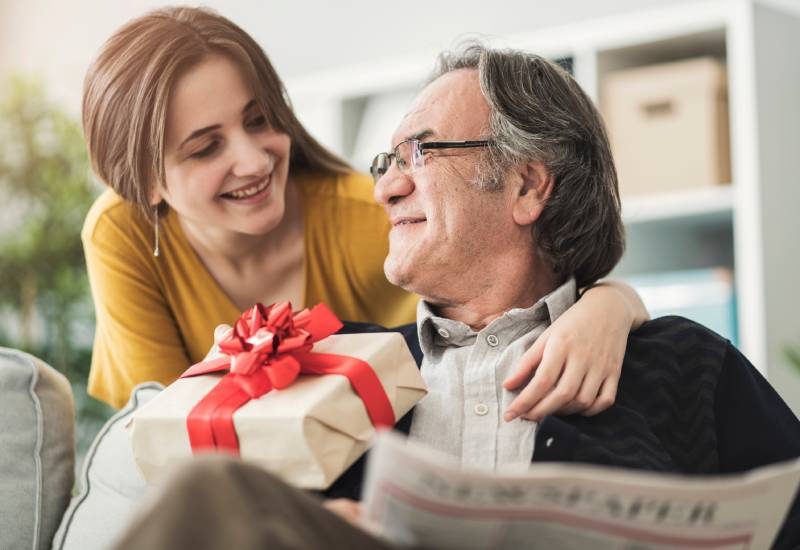 A young woman leaning over the back of a couch and handing a wrapped gift to her dad sitting down with a newspaper in his lap.