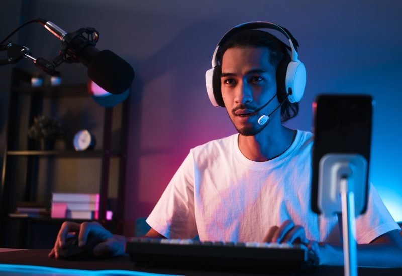 A young man sitting at a desk in a dark room lit by dim purple lights. He is streaming content to an online audience.