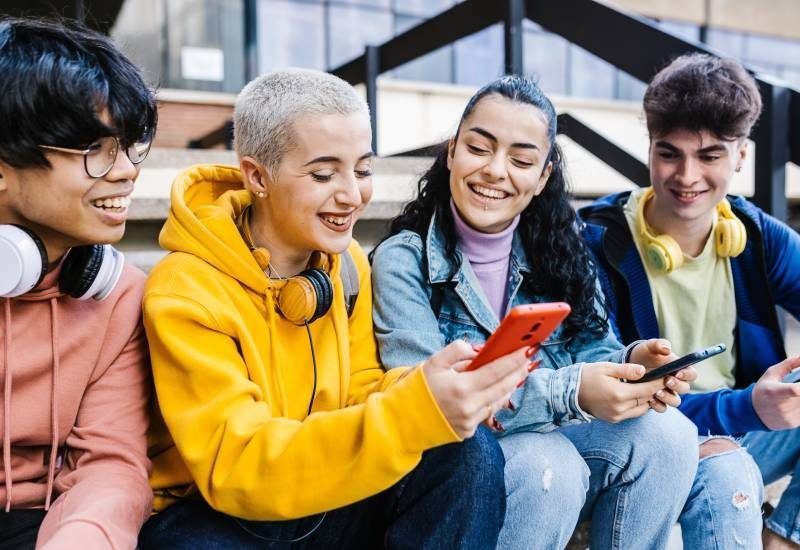 A young girl with short platinum hair shows her friends something on her phone. Everyone is smiling at the phone's contents.