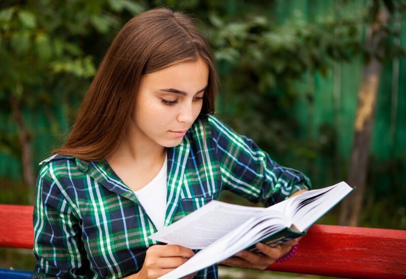 A teenager sitting on a red bench outside is reading a large book. She is holding one page slightly up and waiting to turn it.