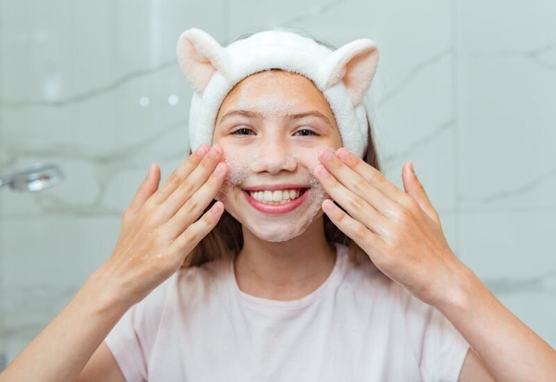 A smiling young girl wearing a white shirt and a white headband rubs cleanser onto her face in a bathroom.