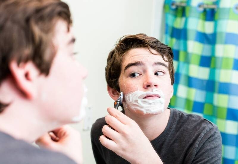 A teenage boy using a disposable razor and shaving cream to shave his face in front of a bathroom mirror.