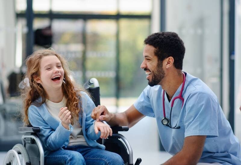 A young girl in a wheelchair smiling as a medical professional in blue scrubs crouches next to her and smiles as well.