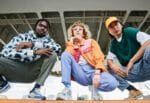 Bugs-eye view of three young adults posing crouched to display their colorful, eccentric outfits under an industrial structure.