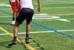 A rear view of a football player stepping through a ladder drill on a turf football field on a sunny day.